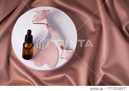 Quartz roller, gua sha and serum bottle on shiny pink fabric. Quartz roller, gua sha and serum bottle on shiny pink fabric. 77701857