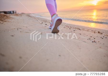 Closeup of runner legs in pink sports shoes performing jog on a sandy beach during sunrise in the early morning 77703266