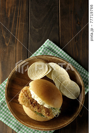 ランチョンマットの上の木皿に乗ったタルタルソースと白身魚のフライサンドとポテトチップスを俯瞰で撮影 77719766