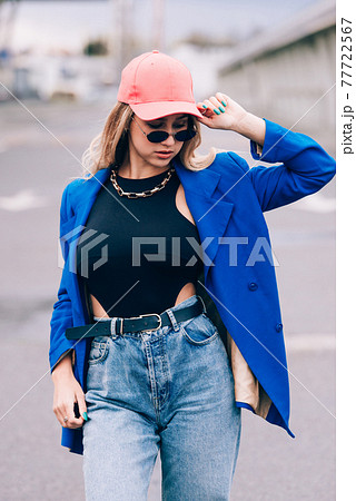 Young sexy blonde hipster woman posing on the street. Wearing blue stylish jacket, jeans and baseball hat and sunglasses. 77722567