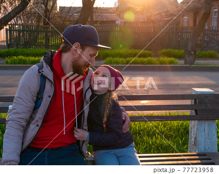 Happy girl with braided hair and father in spring park sits on a bench on sunset outdoor.Kid with dad smiling having fun 77723958