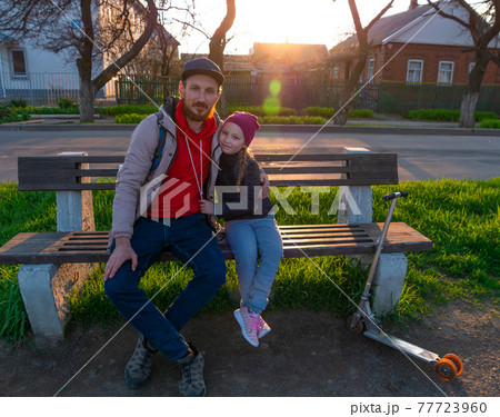 Happy girl with braided hair and father in spring park sits on a bench on sunset outdoor.Kid with dad smiling having fun Happy girl with braided hair and father in spring park sits on a bench on sunset outdoor.Kid with dad smiling having fun 77723960