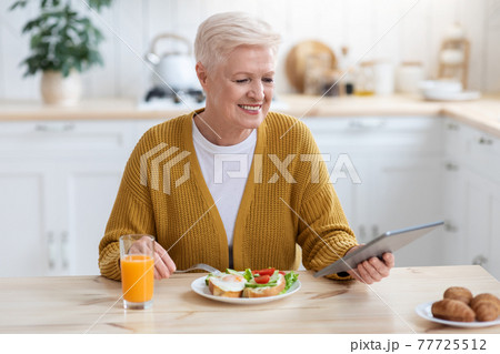 Positive senior woman having lunch and using digital tablet 77725512