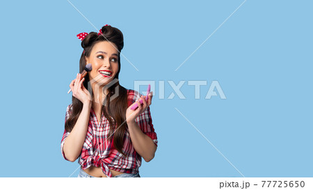Sensual young pinup woman in plaid retro shirt applying decorative makeup with brush on blue studio background Sensual young pinup woman in plaid retro shirt applying decorative makeup with brush on blue studio background 77725650