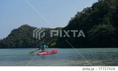 Young blonde woman in blue swimsuit rows pink plastic canoe along azure sea bay past island with palms under blue sky at resort. Young blonde woman in blue swimsuit rows pink plastic canoe along azure sea bay past island with palms under blue sky at resort. 77727428
