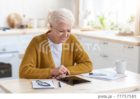 Joyful grey-haired senior woman using digital tablet in kitchen 77757585