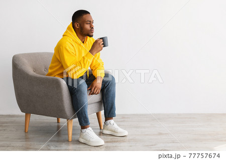 African Guy Thinking Drinking Coffee Sitting Over Gray Background, Side-View African Guy Thinking Drinking Coffee Sitting Over Gray Background, Side-View 77757674