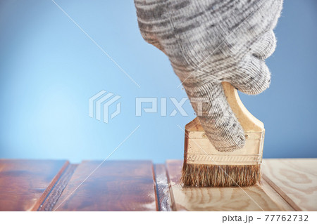 Painting wooden floor with protective varnish on a blue background. Hand in gray work glove painting wood surface. Painting wooden floor with protective varnish on a blue background. Hand in gray work glove painting wood surface. 77762732