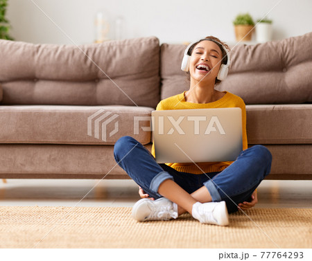 Happy african american teen girl in headphones sitting on floor and listening music while working remotely or studying online 77764293