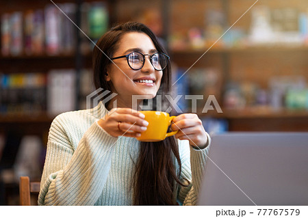 Dreamy positive latin woman relaxing at cafe, enjoying coffee or tea alone, holding mug with hot beverage 77767579