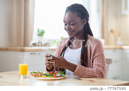 Happy Young Black Female Having Breakfast And Using Smartphone In Kitchen Happy Young Black Female Having Breakfast And Using Smartphone In Kitchen 77767580
