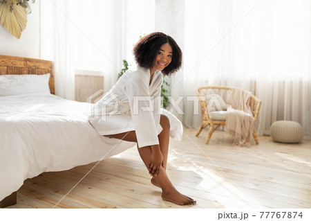 Lovely African American woman in dressing gown touching her leg with silky skin after depilation, sitting on bed at home 77767874