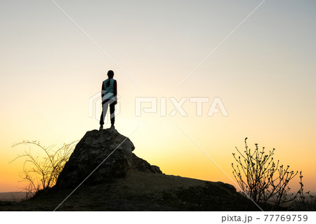 Silhouette of a woman hiker standing alone on big stone at sunset in mountains. Female tourist on high rock in evening nature. Tourism, traveling and healthy lifestyle concept. 77769759