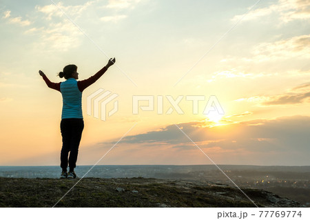 Silhouette of a woman hiker standing alone raising hands enjoying sunset outdoors. Female tourist on rural field in evening nature. Tourism, traveling and healthy lifestyle concept. 77769774