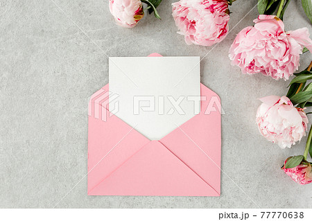 Mockup invitation, blank paper greeting card, pink envelope and peonies on gray stone table. Flower background. Flat lay, top view. 77770638