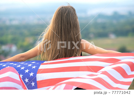 Young happy american woman with long hair holding waving on wind USA national flag on her sholders relaxing outdoors enjoying warm summer day. Young happy american woman with long hair holding waving on wind USA national flag on her sholders relaxing outdoors enjoying warm summer day. 77771042