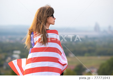 Young pretty american woman with long hair holding waving on wind USA flag on her sholders standing outdoors enjoying warm summer day. Young pretty american woman with long hair holding waving on wind USA flag on her sholders standing outdoors enjoying warm summer day. 77771043