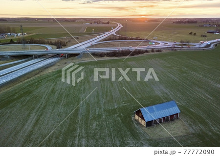 Aerial view of wooden barn in green field on background of modern highway road intersection at dawn on sunny spring day. Drone photography. 77772090