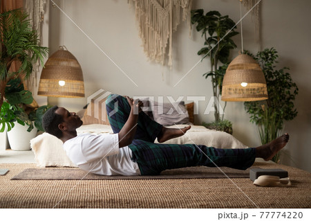 African man doing yoga on the floor, practicing the Supported One Legged Wind Relieving Pose 77774220