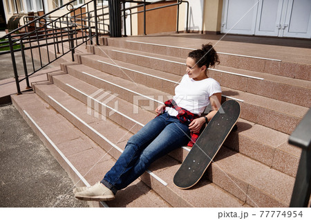 Attractive African American woman in casual clothing posing on stairs with skateboard and enjoying sunny day 77774954