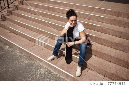 High angle view of a happy smiling woman with skateboard sitting on steps outdoors 77774955