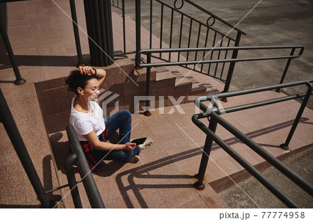 Smiling African American woman sitting on skateboard with headphones and smartphone in her hand and enjoying beautiful sunny day Smiling African American woman sitting on skateboard with headphones and smartphone in her hand and enjoying beautiful sunny day 77774958