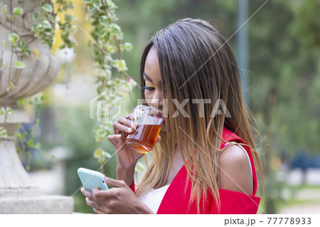Beautiful young African girl with a Cup of tea and a phone in her hands in the Park Beautiful young African girl with a Cup of tea and a phone in her hands in the Park 77778933
