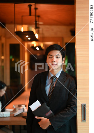 A businessman is holding a document file while standing over the meeting room as a background. 77790268