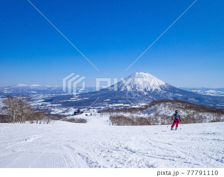 快晴の日に羊蹄山を望む閑静なゲレンデを滑走するスキーヤー (ニセコ、北海道) 77791110