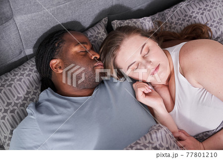 Multiracial couple laying at the bed indoors at home and hugging while sleeping Multiracial couple laying at the bed indoors at home and hugging while sleeping 77801210