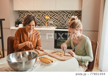 Girl chopping with the knife fresh banana while baking bagels with her grandmother 77801404