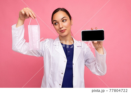 pretty female doctor wearing white coat holding mask showing modern mobile phone isolated over pink 77804722