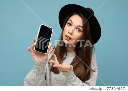 Portrait photo shot of beautiful young woman wearing black hat and grey sweater holding phone 77804907