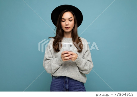 Beautiful young brunette woman wearing black hat and grey sweater holding smartphone looking down at 77804915