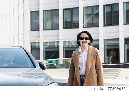 A beautiful confident woman is standing near a car on the background of a business center and smiling. A beautiful confident woman is standing near a car on the background of a business center and smiling. 77809082