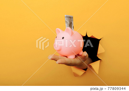Hand with a piggy bank and dollar banknote through a yellow paper hole. Investment and savings Hand with a piggy bank and dollar banknote through a yellow paper hole. Investment and savings 77809344