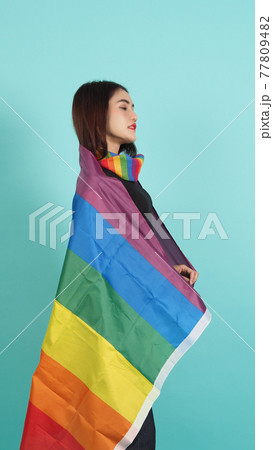 LGBTQ girl and pride flag. Sexy Lesbian girl and LGBT flag standing. blue green background. Asian LGBTQ woman with rainbow scarf on neck. Energetic cheerful. LGBTQ concept.  77809482