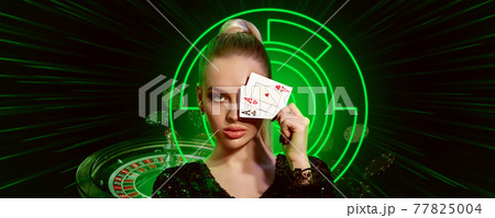 Woman in black dress. Covered eye by two aces, posing on black background with neon lights, roulette and falling chips. Poker, casino. Close-up 77825004