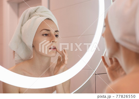 Woman standing near mirror in the bathroom and applying moisturizer her face 77826550