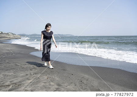 海風に吹かれながら砂浜の波打ち際を歩く若い女性（神奈川県鎌倉市七里ヶ浜） 77827972