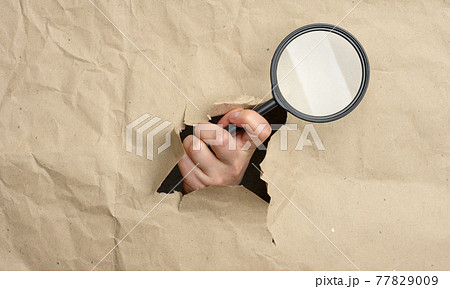 a woman's hand holds a glass magnifier, part of the body sticks out of a hole in brown paper 77829009
