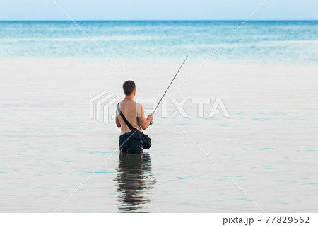 A young guy without a shirt stands knee-deep in the water of the sea in the evening and catches fish with a line 77829562