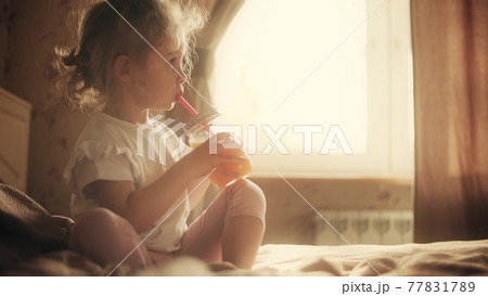 Cute blonde little girl drinks orange juice with a straw at home 77831789