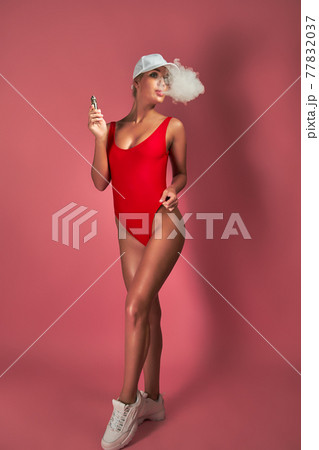 Seductive glamour young woman in red swimsuit standing and vaping on pink background 77832037