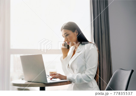 Beautiful woman talking on phone while  working on laptop computer sitting at home managing her business via home office during Coronavirus or Covid-19 quarantine 77835537