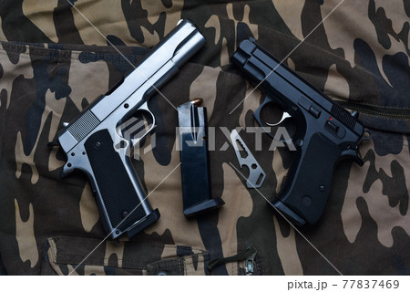 Still life with black and silver handgun with cartridge magazine and pocket knife tool on camouflage soldier pant background, Black and chrome semi-automatic pistol on camouflage soldier pant Still life with black and silver handgun with cartridge magazine and pocket knife tool on camouflage soldier pant background, Black and chrome semi-automatic pistol on camouflage soldier pant 77837469