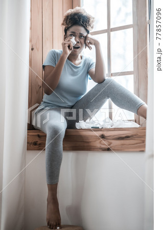 Crying woman sitting at home on windowsill 77857096