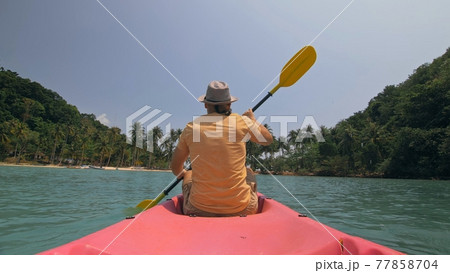 Man with sunglasses and hat rows pink plastic canoe along sea against green hilly islands with wild jungles. Traveling to tropical countries. Man with sunglasses and hat rows pink plastic canoe along sea against green hilly islands with wild jungles. Traveling to tropical countries. 77858704