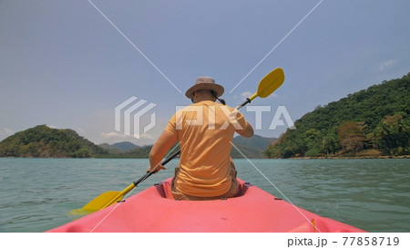 Man with sunglasses and hat rows pink plastic canoe along sea against green hilly islands with wild jungles. Traveling to tropical countries. 77858719