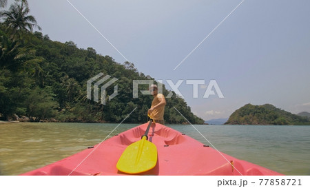 Man with sunglasses and hat rows pink plastic canoe along sea against green hilly islands with wild jungles. Traveling to tropical countries. 77858721